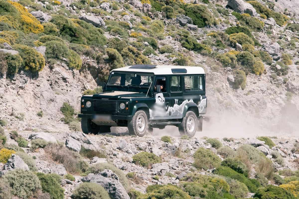 Georgioupolis land rover safari: a dark green Land Rover Defender with a white roof drives along a dusty, rocky track on a sunlit mountain slope in Crete, with yellow broom bushes, grey limestone rocks and a trail of pale dust around it.