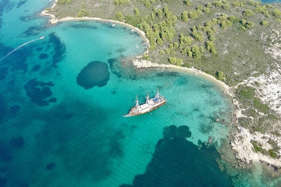 Halkidiki boat trip: an aerial view of a three-masted pirate-style wooden ship at anchor in clear turquoise water beside a sandy-edged, pine-dotted headland on a Blue Lagoon cruise, with small boats in the bay.