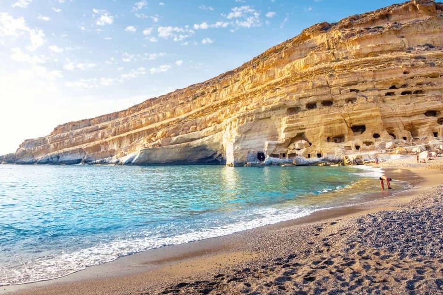 Heraklion beach trip: a tall golden sandstone cliff with rows of dark carved cave openings rises above a curved pebble beach and clear turquoise water with small white waves, under a pale blue sky.
