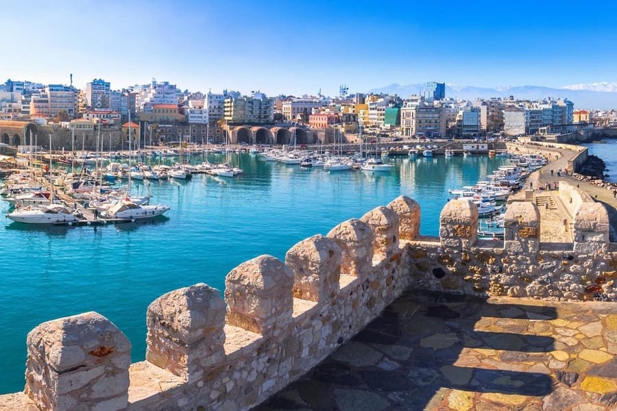 Heraklion castle tour: the crenellated stone battlement of a sea fortress in the foreground looks over a turquoise harbour full of white yachts, with a row of brick-red vaulted arches on the quay, pastel town buildings and pale mountains on the horizon under a bright blue sky.