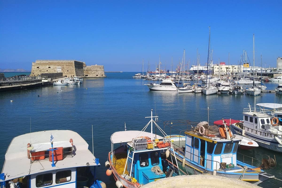 Heraklion day trip: a crenellated stone sea fortress stands at the end of a harbour of calm deep blue water, with white yachts and sailing boats moored beyond it and blue-and-white fishing boats in the foreground under a clear blue sky.
