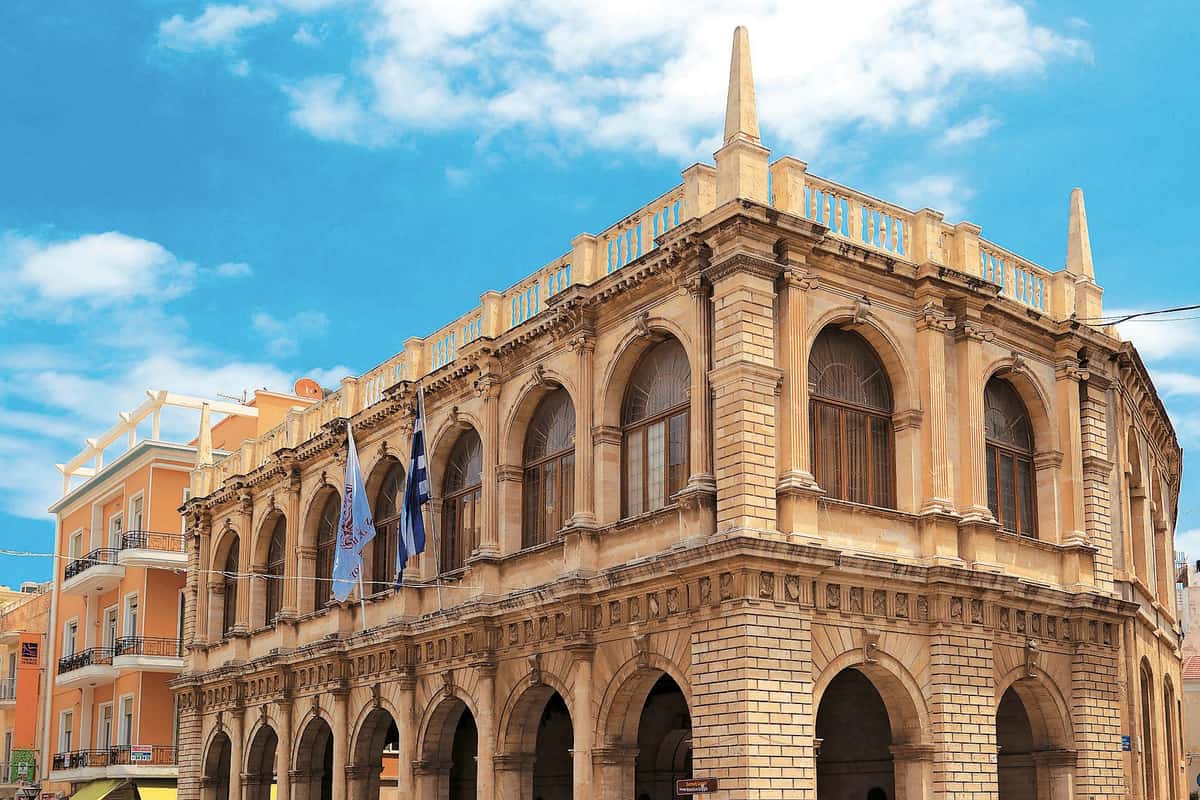 Heraklion walking tour: a large sandstone building with arched windows on the upper floor and an open arcade of arches below, a stone balustrade with pointed spires, a blue-and-white flag on the corner and an orange building beside it, under a bright blue sky with white clouds.