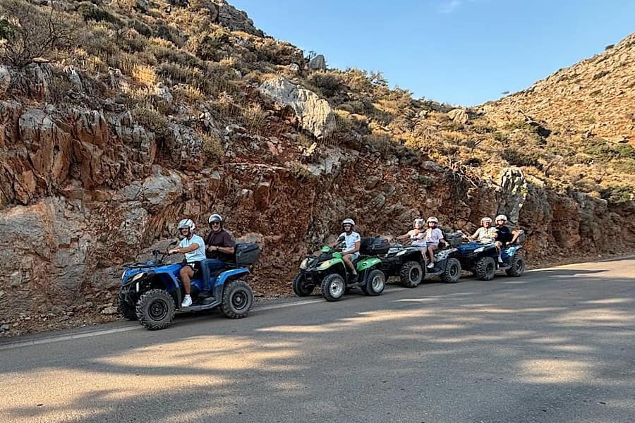 Hersonissos ATV tour: a line of blue, green and black quad bikes with helmeted riders stands on a paved mountain road below a red and grey rock cut covered in dry scrub, under a clear blue sky.