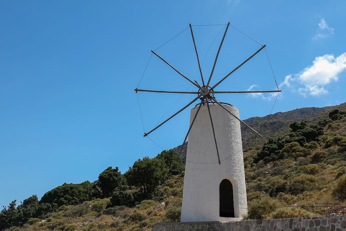 Hersonissos ATV tour: a white round Lasithi windmill tower with an arched doorway and a wooden sail frame stands on a green scrubby hillside below a deep blue sky with a few white clouds.