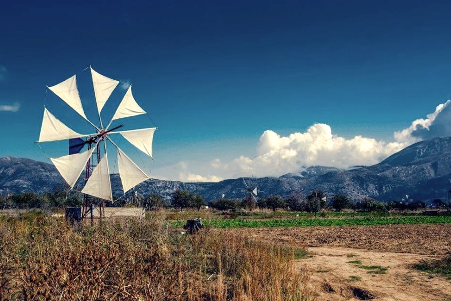 Hersonissos jeep safari: a white sail windmill on a steel frame stands beside a dirt track and dry grass on the Lasithi Plateau, with more windmills in the flat fields, a mountain range and a big white cloud under a deep blue sky.