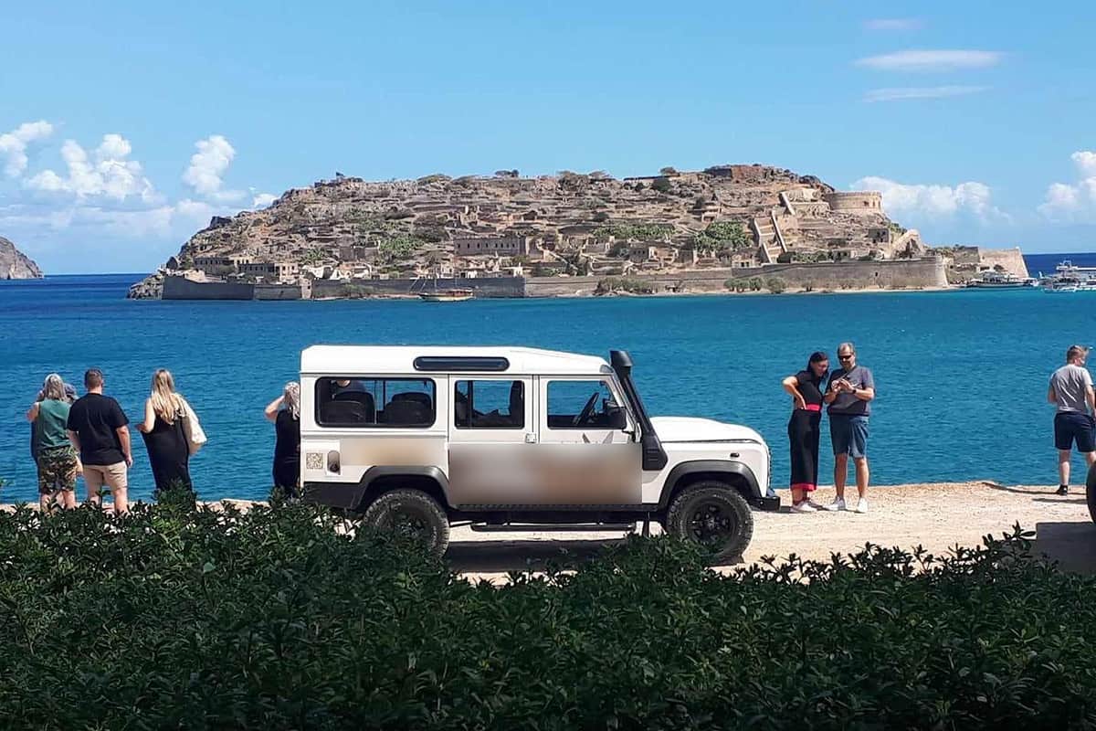 Hersonissos jeep safari: a white Land Rover 4x4 with a snorkel stands on a gravel viewpoint facing Spinalonga island, whose old fortress walls and ruined stone buildings rise from deep blue water under a bright blue sky, with a few visitors looking at the view and a green hedge in front.