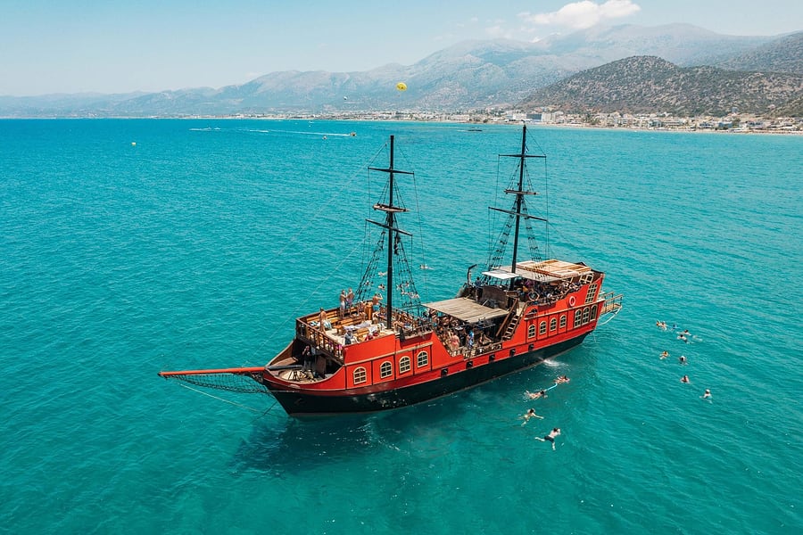 Hersonissos transfer: an aerial view of a red and black wooden pirate ship with two tall masts anchored in turquoise water off the Hersonissos coast, with swimmers nearby and a beach town below the mountains in the distance.