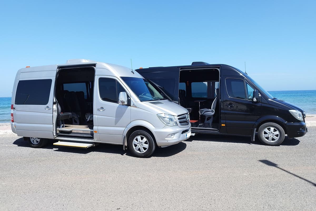 Hersonissos transfer: a silver minibus and a black minibus with their sliding doors open stand side by side on a coastal road beside the blue sea under a clear blue sky.
