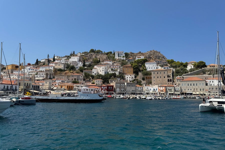 Hydra boat trip: a harbour town seen from the water, with stone mansions and terracotta roofs below a rocky hill, small boats moored along the quay, a clock tower and sailing yachts in the foreground under a clear blue sky.