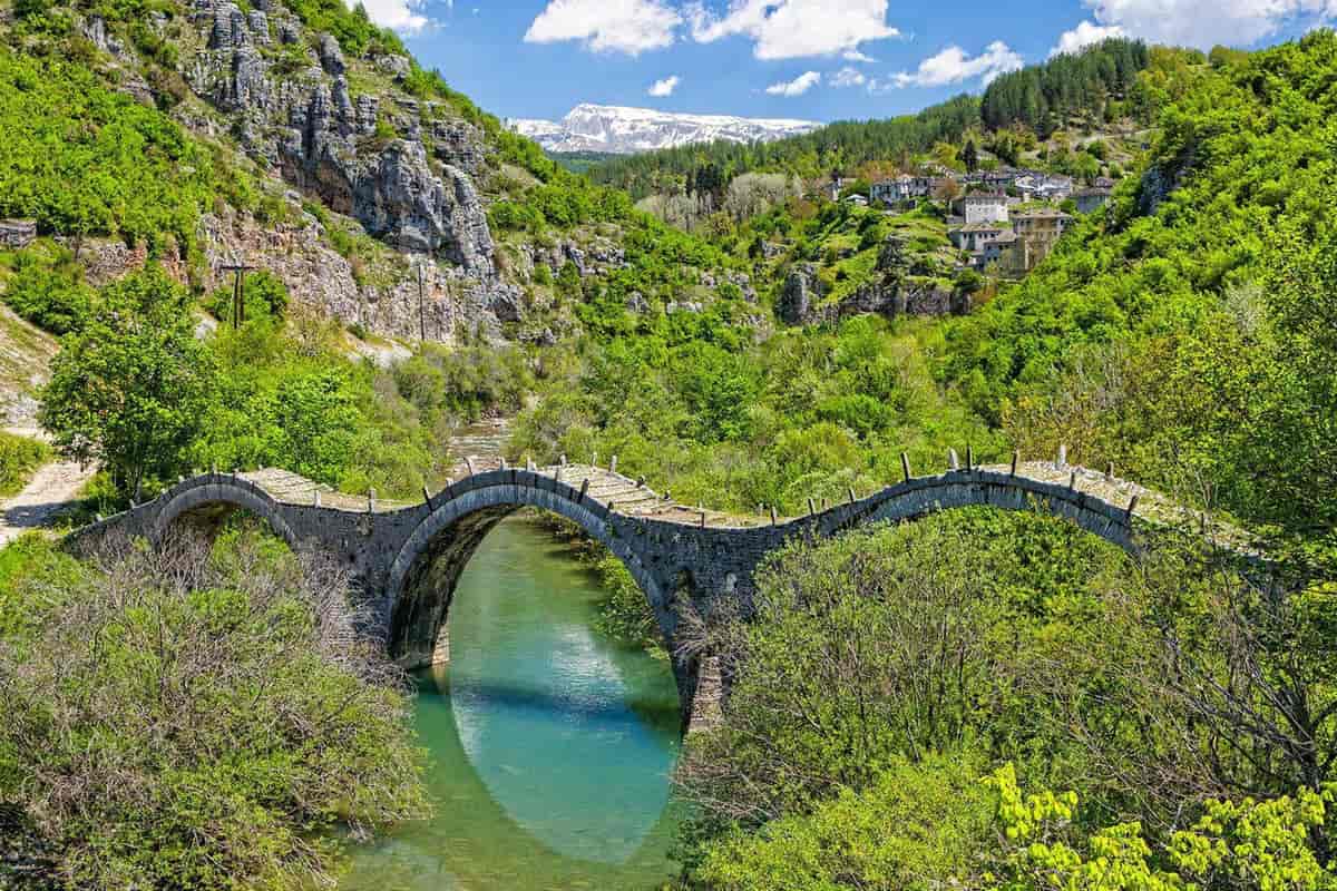 Ioannina Zagori tour: the three-arch stone Kalogeriko bridge crosses a turquoise river in a green gorge, with Zagori stone houses on the hillside and a snow-capped mountain under a blue sky.