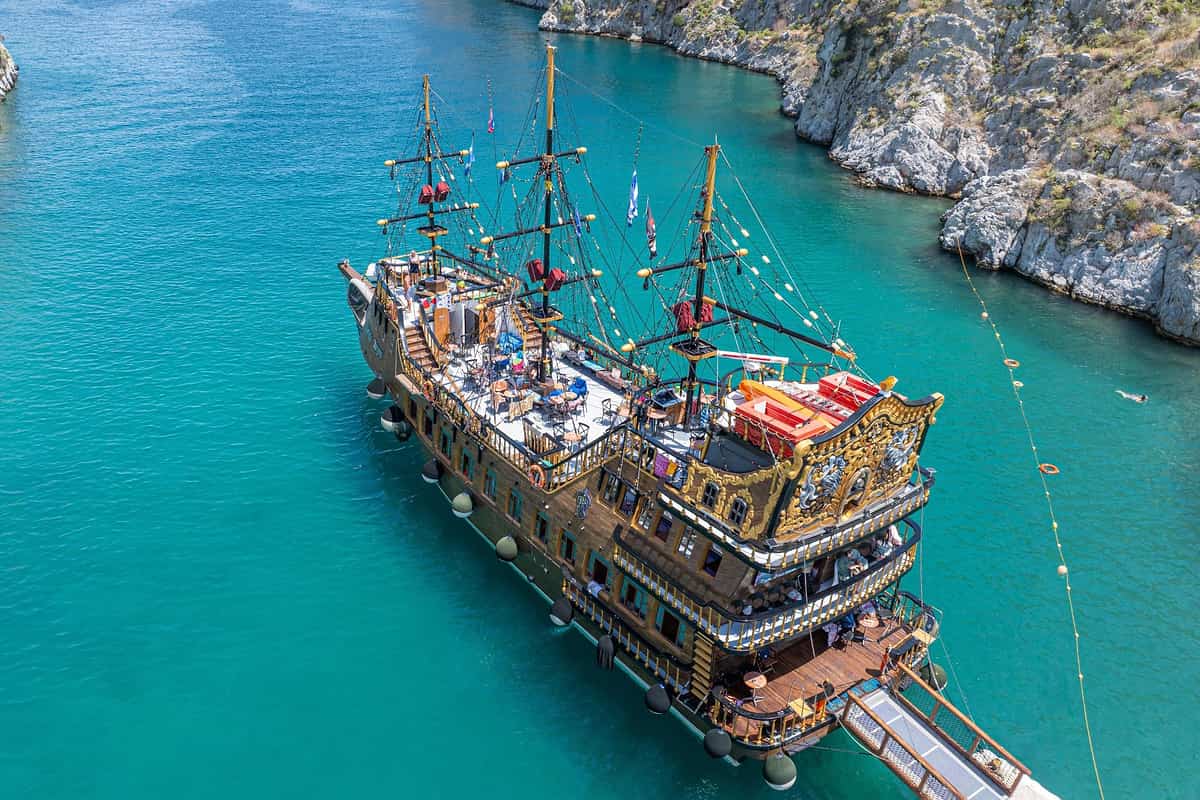 Kalymnos boat trip: a three-masted wooden pirate-style cruise ship with a gold-trimmed stern and flags lies moored beside a stone pier in a narrow turquoise channel between pale rocky cliffs, seen from above.