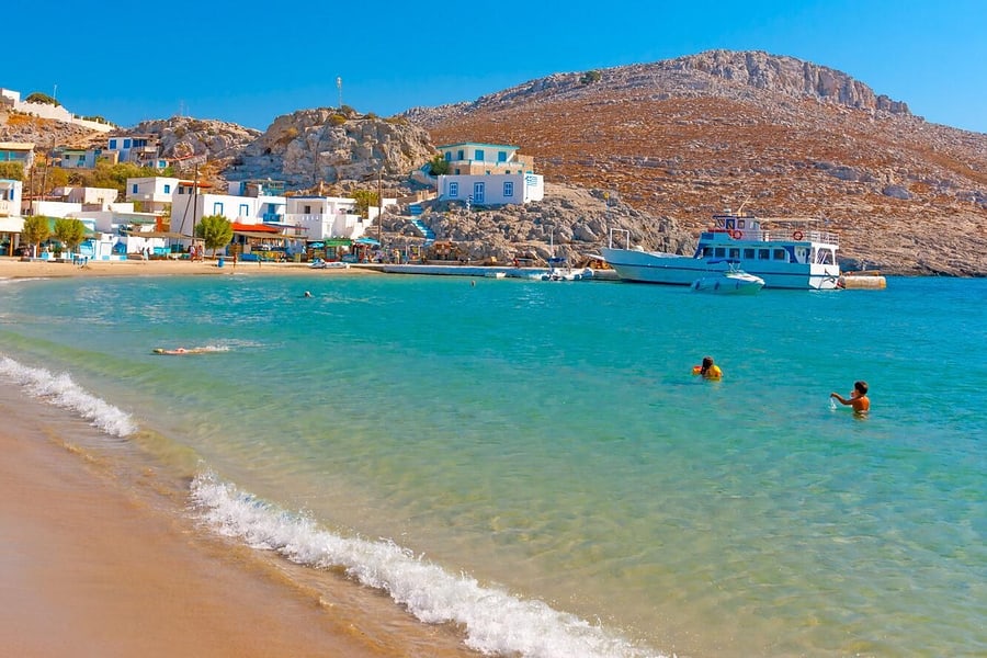 Kalymnos boat trip: a curved sandy beach with clear turquoise shallows and a white wave, white and blue houses below a bare rocky hill, a white passenger boat moored at the quay and two swimmers in the water.