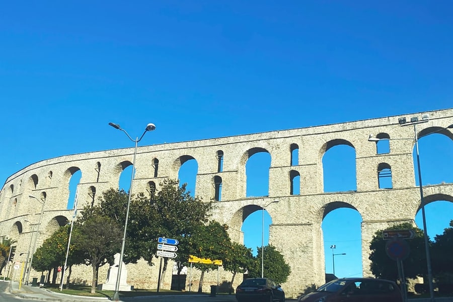 Kavala day trip: a long curved stone aqueduct with two tiers of tall arches rises behind orange trees and street lamps under a bright blue sky.