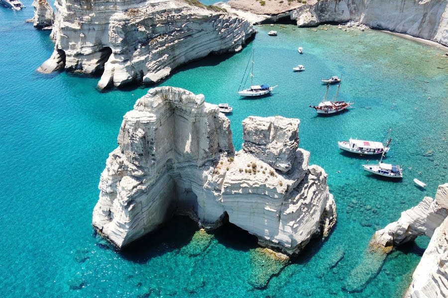 Kleftiko boat tour: an aerial view of two tall white rock pillars with a sea arch rising from clear turquoise water, with sailboats, a wooden two-masted boat and small boats anchored in the bay below white cliffs.