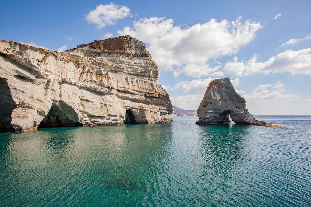 Kleftiko boat tour: a tall layered white and cream cliff with dark sea cave openings and a smaller rock arch stand in calm green-turquoise water under a blue sky with white clouds, with a hazy coastline in the distance.