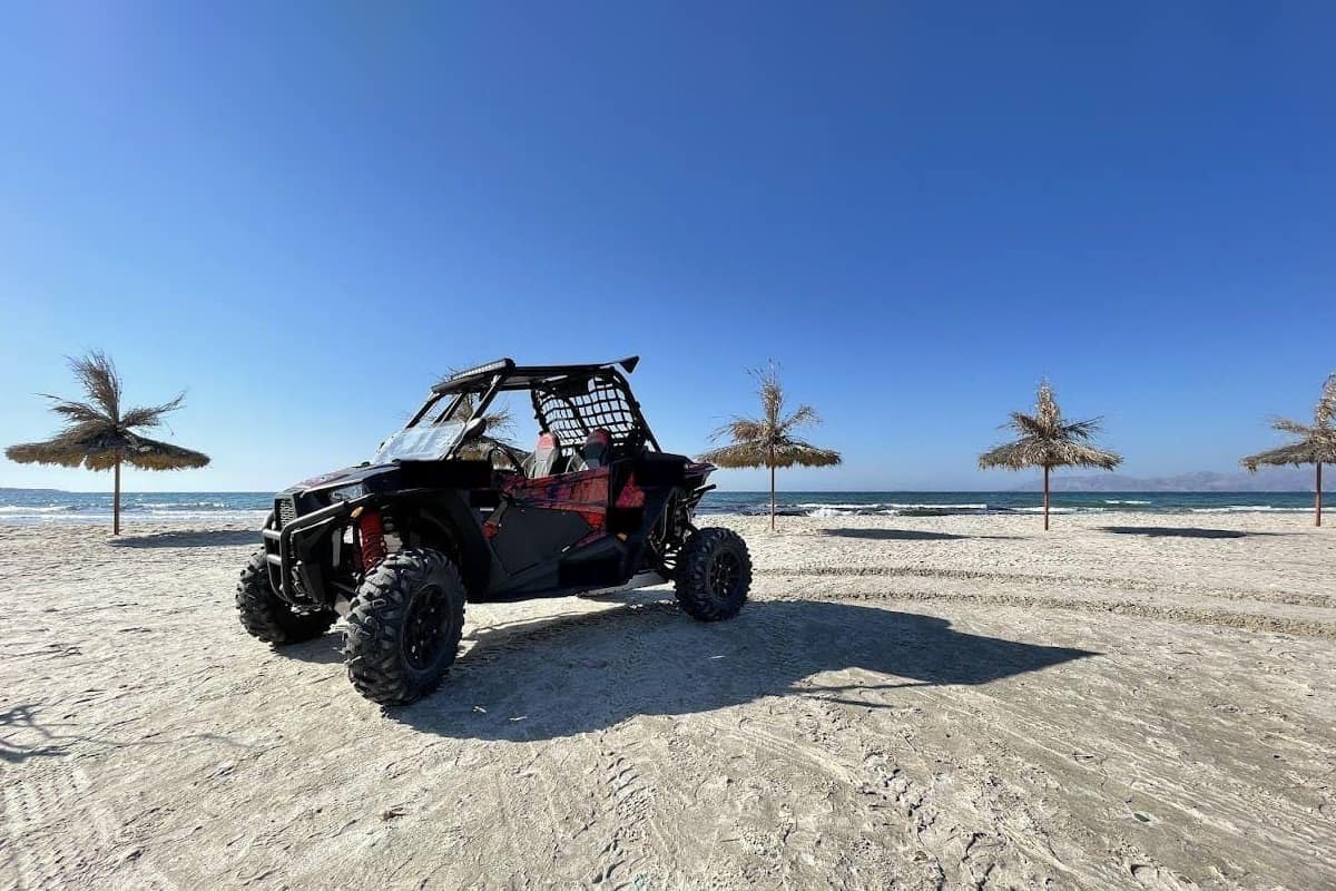 Kos ATV tour: a black and red off-road buggy stands on pale sand beside straw beach sunshades and a calm blue sea on the coast of Kos, under a clear deep blue sky.