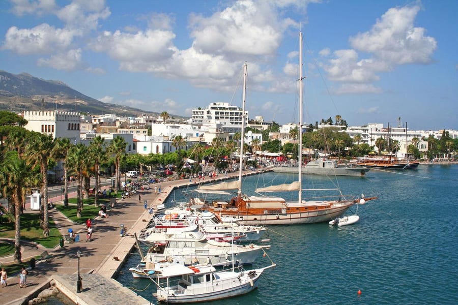Kos ATV tour: a wooden sailing yacht and white motor boats moored along the palm-lined waterfront promenade of Kos Town harbour, with white buildings and a mountain ridge behind under a blue sky with white clouds.