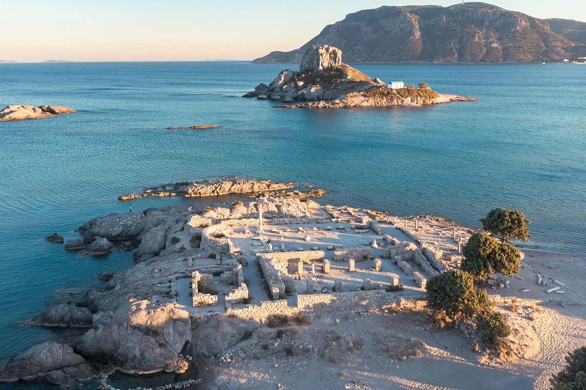 Kos beach trip: an aerial view of a rocky seaside point with low stone ruins and a few trees beside a sandy shore, a small rocky islet with a white chapel in a calm blue bay and a large mountain across the water in warm evening light.