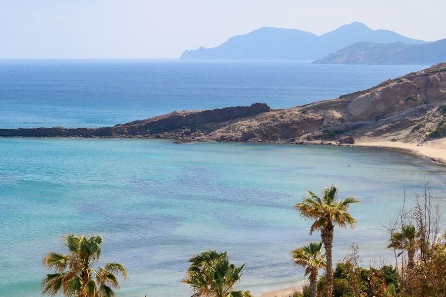 Kos beach trip: a wide turquoise bay with a rocky headland and a pale sandy beach curving along the shore, palm trees in the foreground and blue hills on the horizon under a hazy sky.