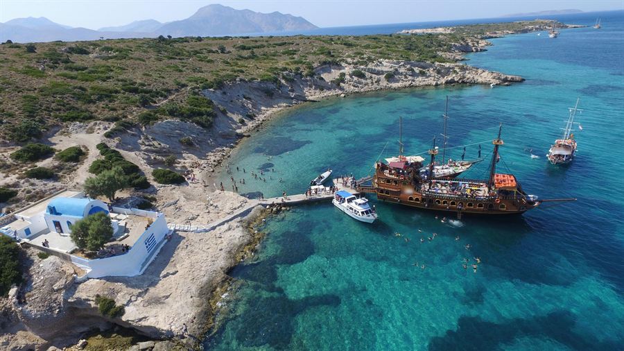 Kos boat trip swim stop: a pirate-style cruise ship moored at a small islet with a white chapel and swimmers