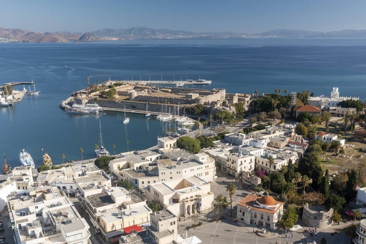 Kos day trip: aerial view of a stone castle on a harbour peninsula with moored yachts, a town square and white buildings beside the blue sea.