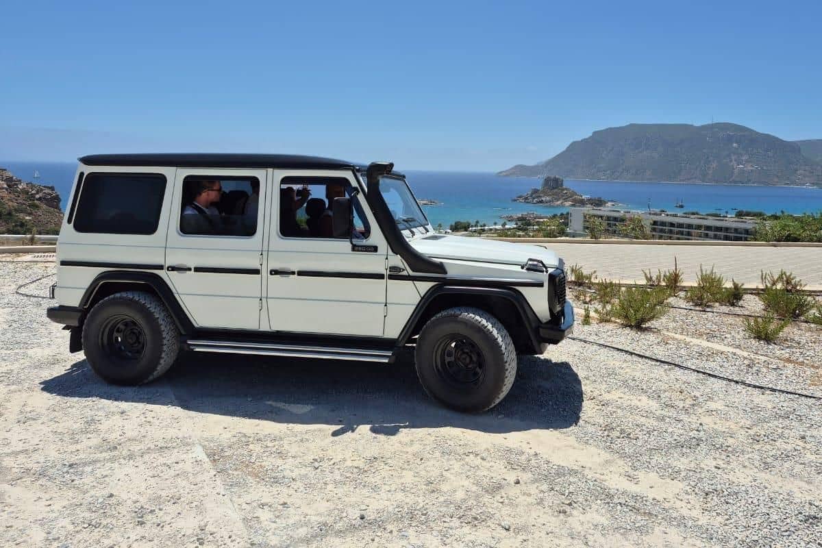 Kos jeep safari: a white Mercedes G-Class 4x4 with a black roof and snorkel stands on a gravel viewpoint above the blue bay of Kefalos, with the rocky islet of Kastri and its small chapel, a hotel on the shore and a large mountain across the sea under a clear blue sky.