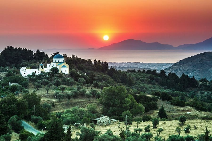 Kos jeep safari: the sun sets in an orange-red sky over the sea and distant island mountains, above a white church with a blue dome and a yellow side chapel on an olive-covered hillside near Zia on the slope of Mount Dikeos, with a winding road below.