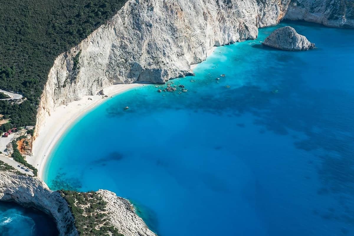 Lefkada beach trip: an aerial view of a curved white beach below tall pale cliffs, with bright turquoise water fading into deep blue sea, a large rock standing in the bay and a dark green forested hillside above.