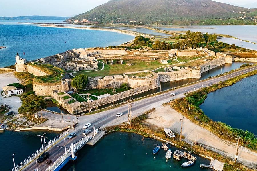 Lefkada private tour: an aerial view of a large stone fortress with moats, a small lighthouse and green courtyards beside a lagoon, with a road and a bridge crossing the water and a green mountain behind.