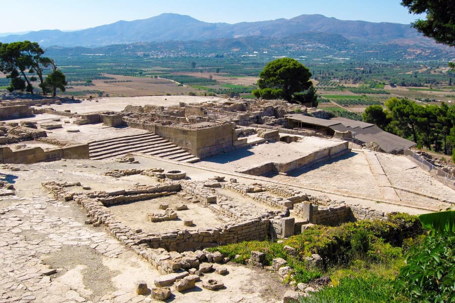 Matala day trip: low stone walls, paved courts and a wide stone staircase of an ancient hilltop palace site look out over a green farmland plain and blue mountains, with a pine tree and a covered shelter beside the ruins.