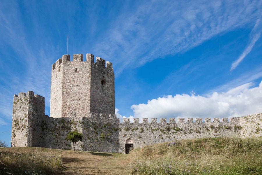 Meteora castle tour: a tall crenellated stone tower and a long battlemented stone wall stand on a grassy hill under a deep blue sky with white clouds.