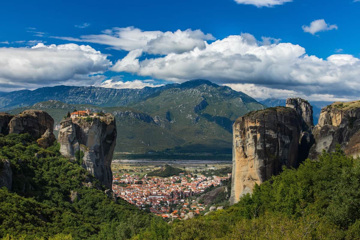 Meteora day trip: a stone monastery with red roofs stands on top of a tall, narrow rock pillar on the left, a huge rock cliff rises on the right, and between them the red-roofed houses of a town fill the green valley below mountains and a blue sky with white clouds.
