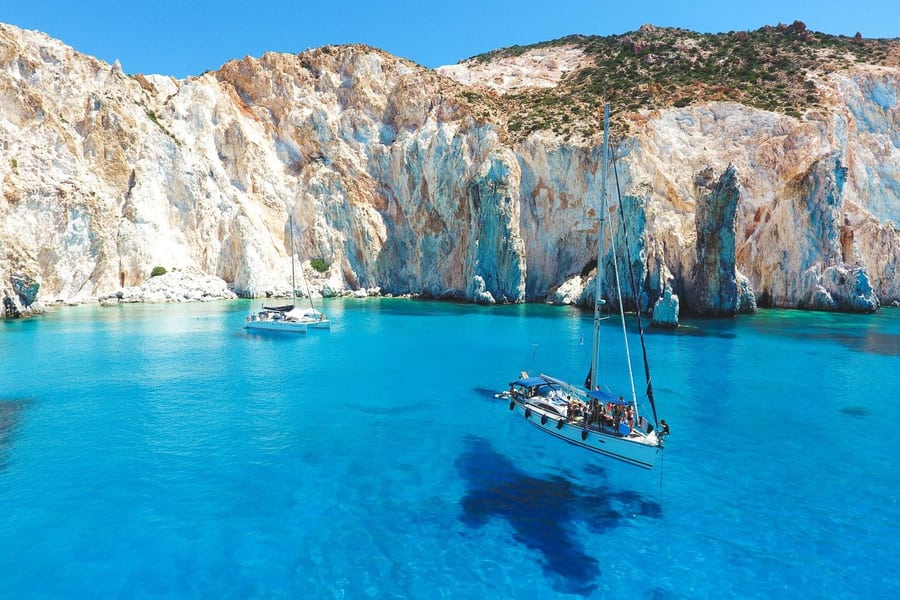 Milos day trip: a sailboat and a catamaran lie on bright turquoise water below tall white and ochre cliffs with rock spires and green scrub on top, under a deep blue sky.