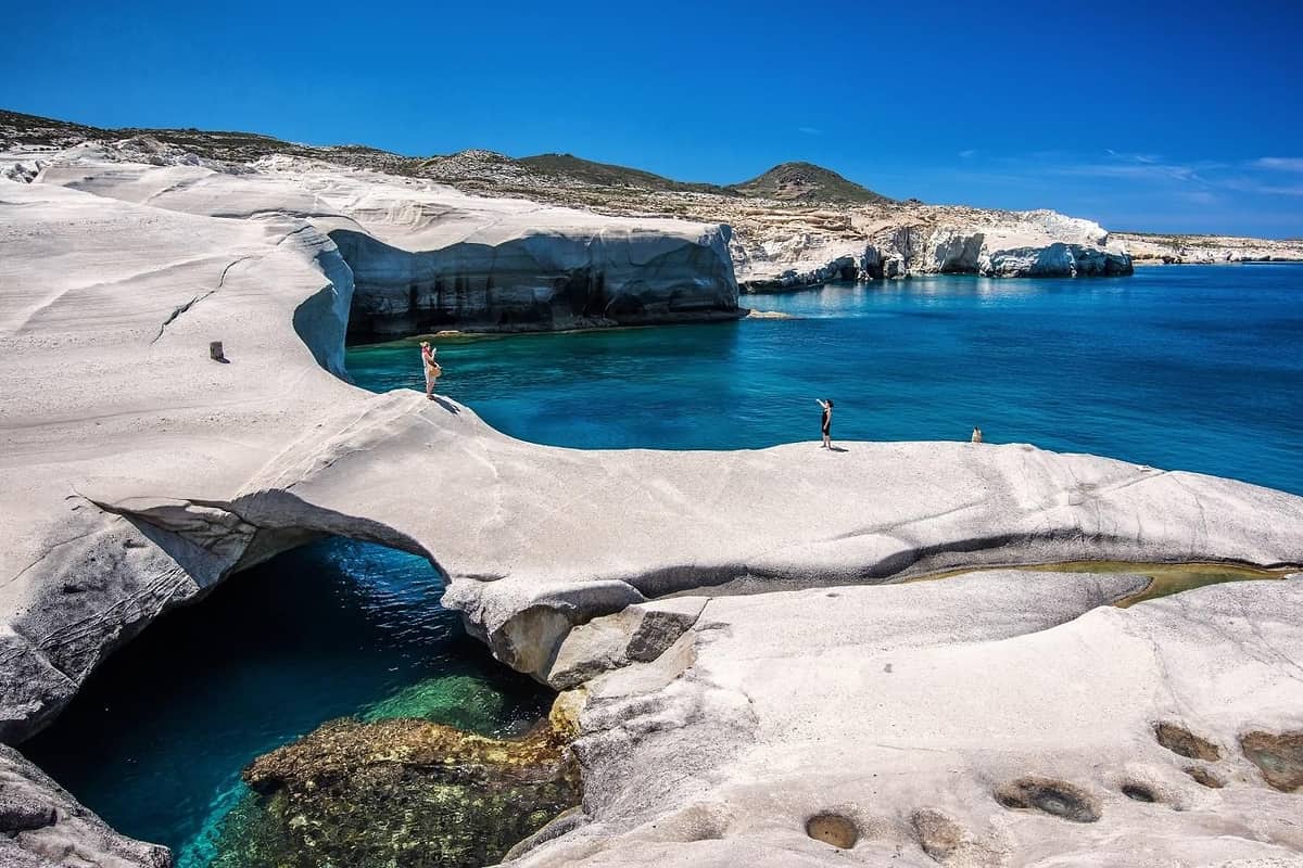 Milos day trip: smooth white volcanic rock curves around a deep blue sea inlet with a dark water-filled arch below, two small walkers on the rock and a low hill under a clear blue sky.
