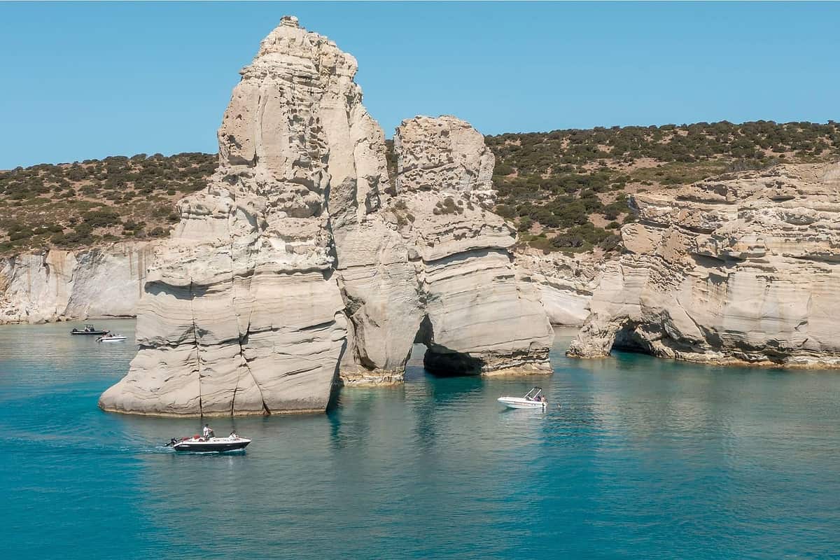 Milos private tour: a tall pale rock pillar with a smaller rock beside it rises from calm turquoise water below low cliffs and scrubby hills, with a few small boats on the bay under a clear blue sky.