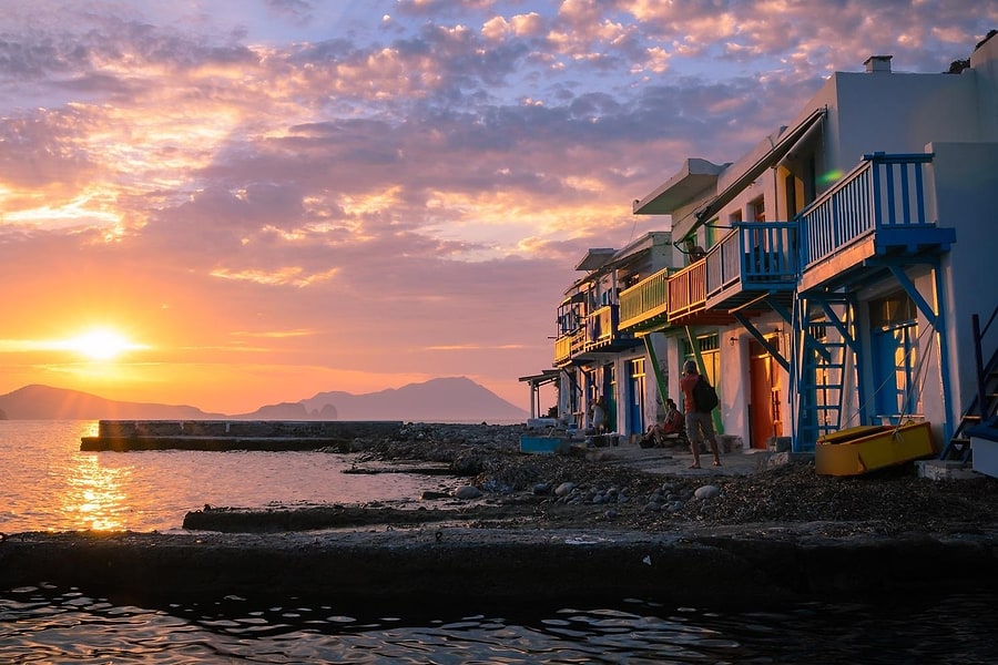 Milos private tour: a row of white harbour houses with blue, green and orange wooden balconies stands at the water's edge at sunset, with the sun low behind distant island silhouettes under a pink and purple cloudy sky.
