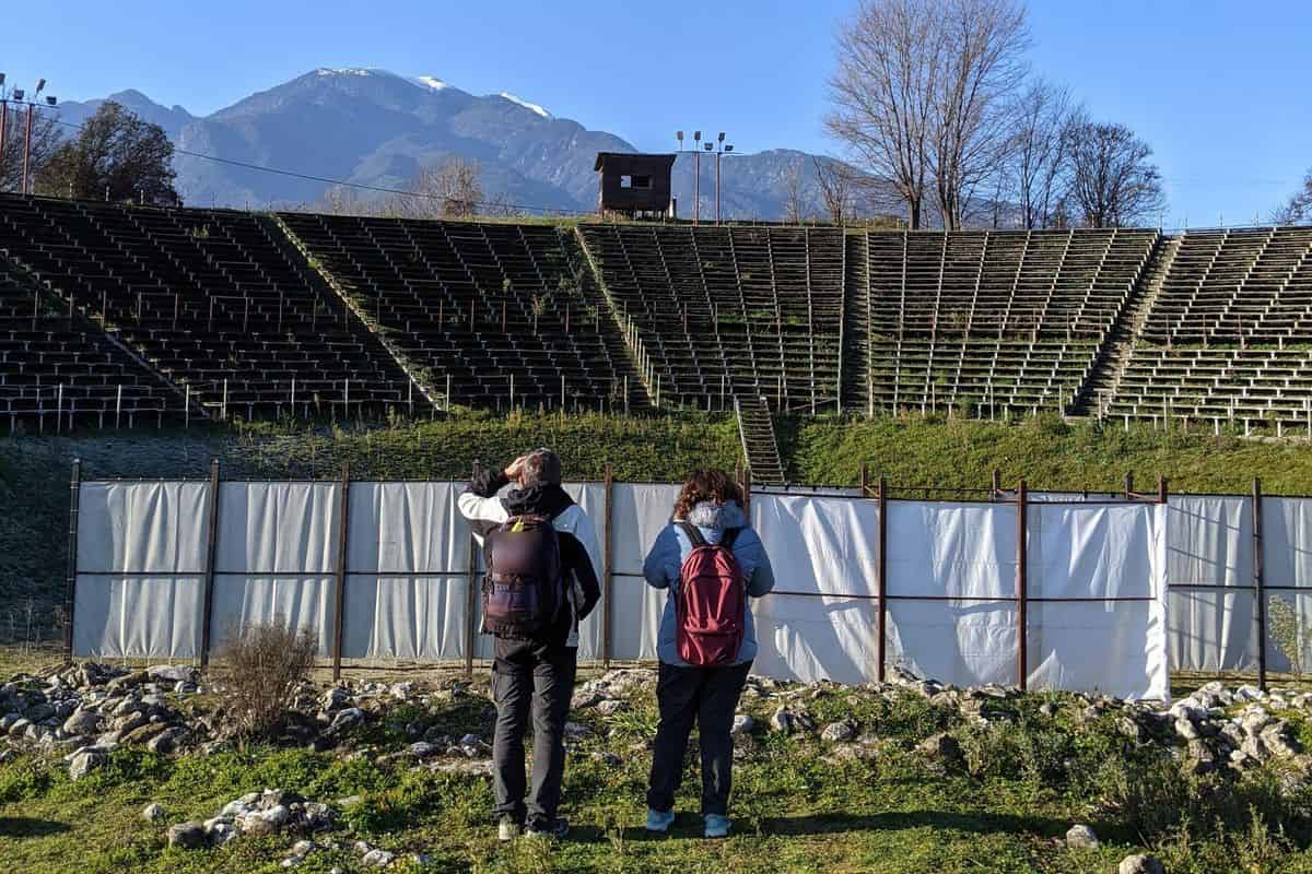 Mount Olympus day trip: two visitors in backpacks look up at the snowy peak above the ancient theatre at Dion