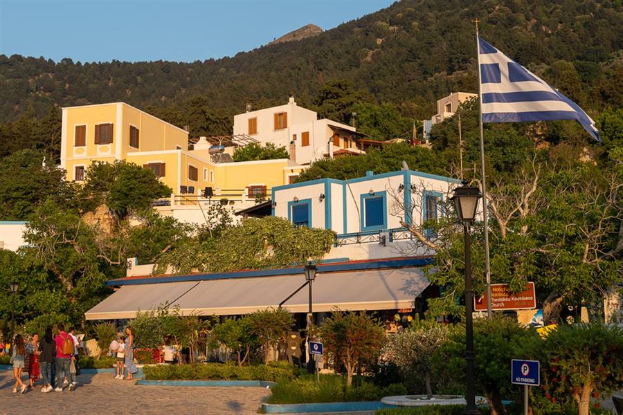Mount Zia sunset: yellow and whitewashed houses of Zia village climb a wooded mountain slope above a paved square, where visitors walk past a Greek flag in warm evening light.
