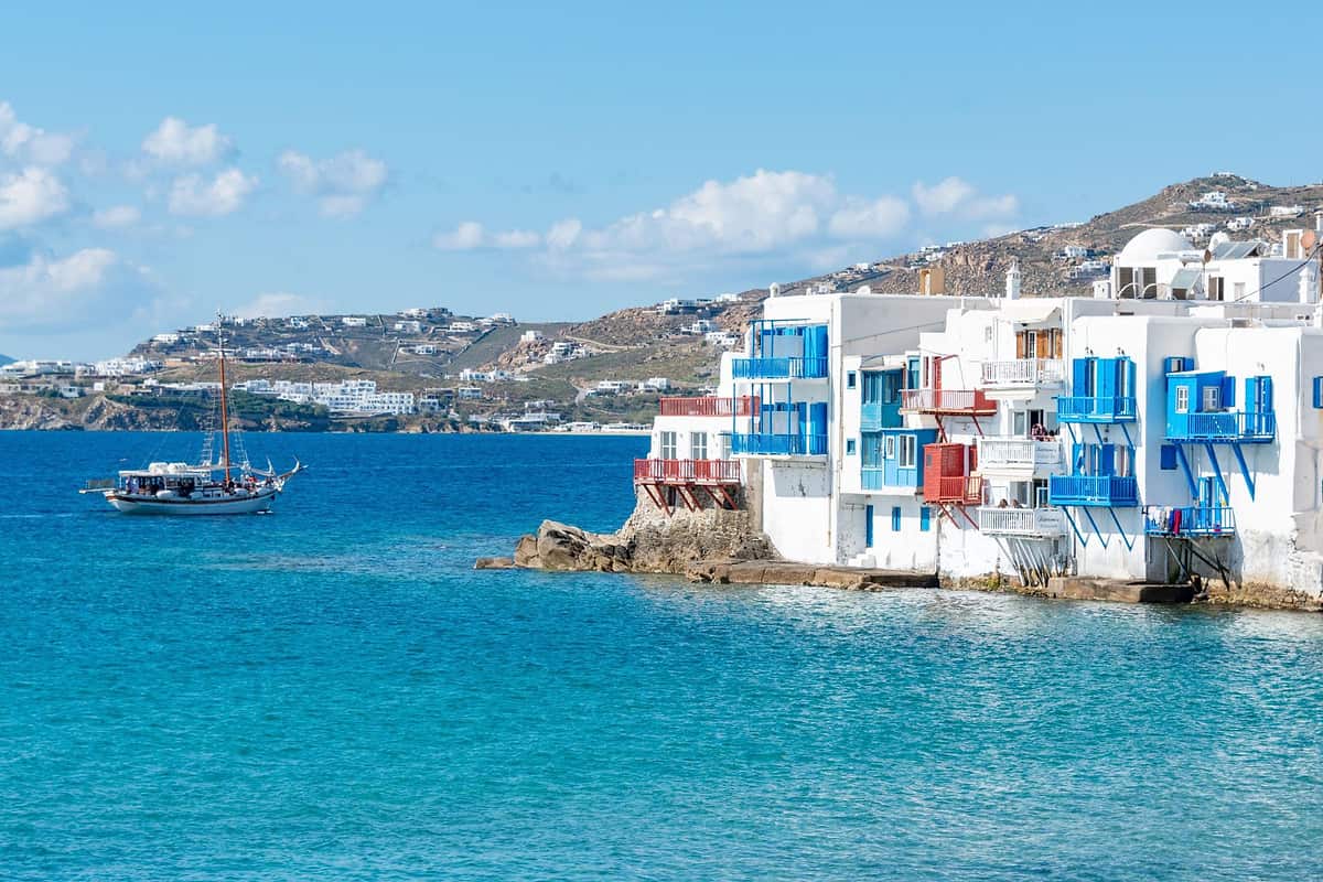 Mykonos boat trip: a wooden two-masted sailing boat with passengers on deck cruises across bright blue water towards the white houses of Little Venice, whose blue and red wooden balconies hang over the sea, with brown hills and a few white clouds behind under a clear blue sky.