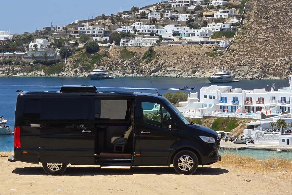 Mykonos transfer: a black minibus with its sliding door open stands on a sandy hilltop above a deep blue bay, with whitewashed buildings with blue and red shutters, yachts on the water and a hillside of white houses behind it.