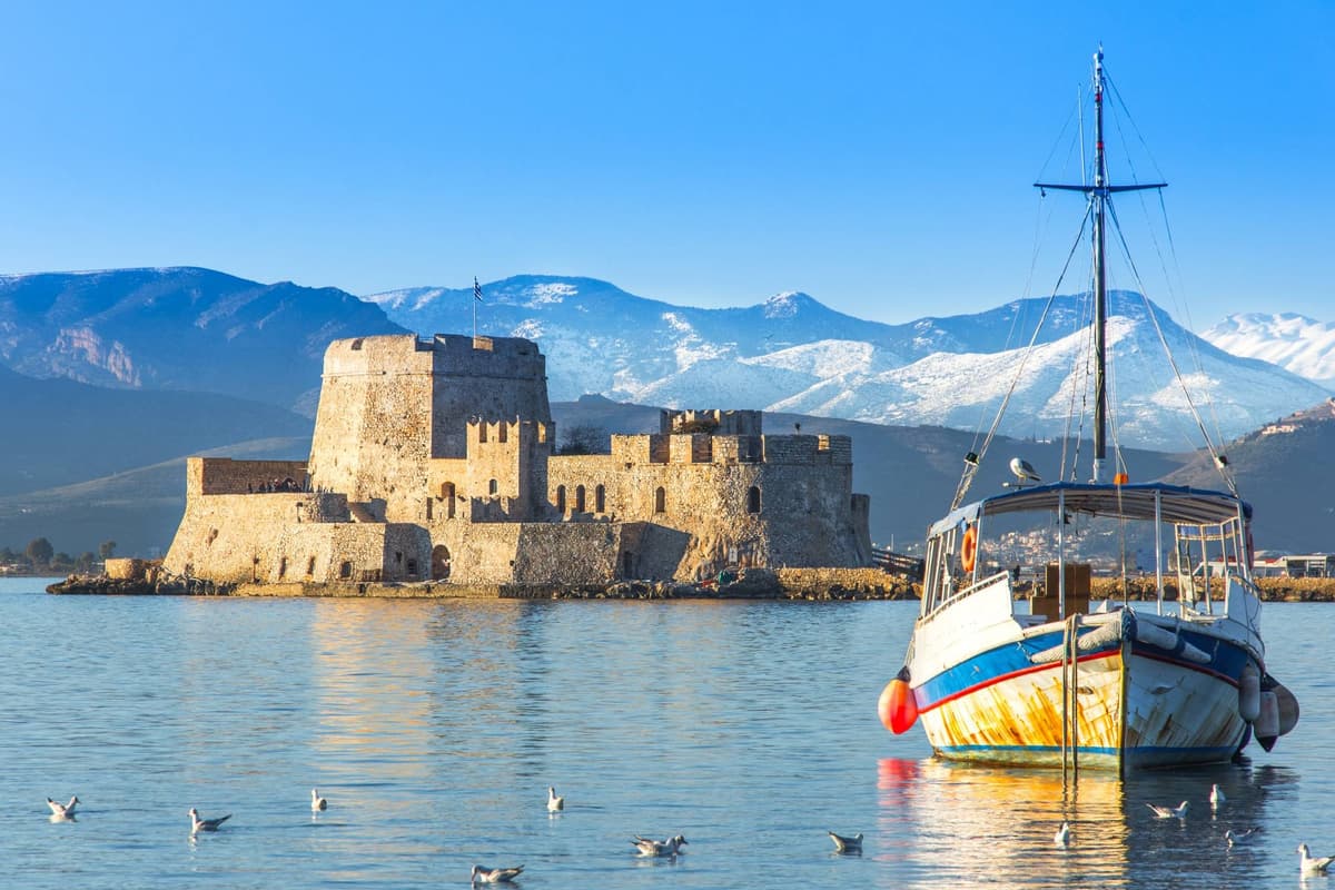 Nafplio day trip: the golden-stone Bourtzi sea castle stands on its small islet in the calm bay of Nafplio in warm sunlight, with a colourful old fishing boat and seagulls on the water and snow-capped mountains behind under a clear blue sky.