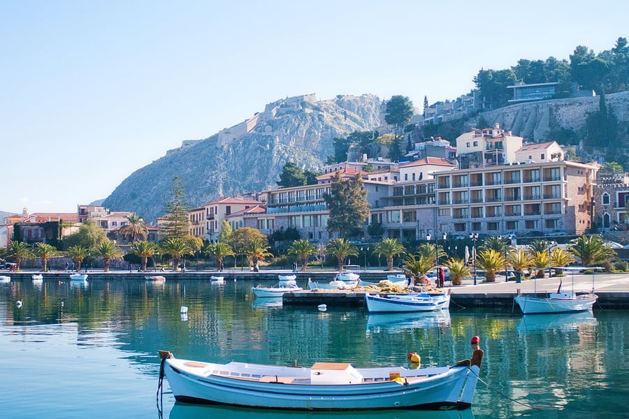 Nafplio day trip: the Palamidi fortress crowns a steep rocky hill above the old town and the harbour promenade of Nafplio, with palm trees, pastel buildings and white fishing boats moored in the calm turquoise water.