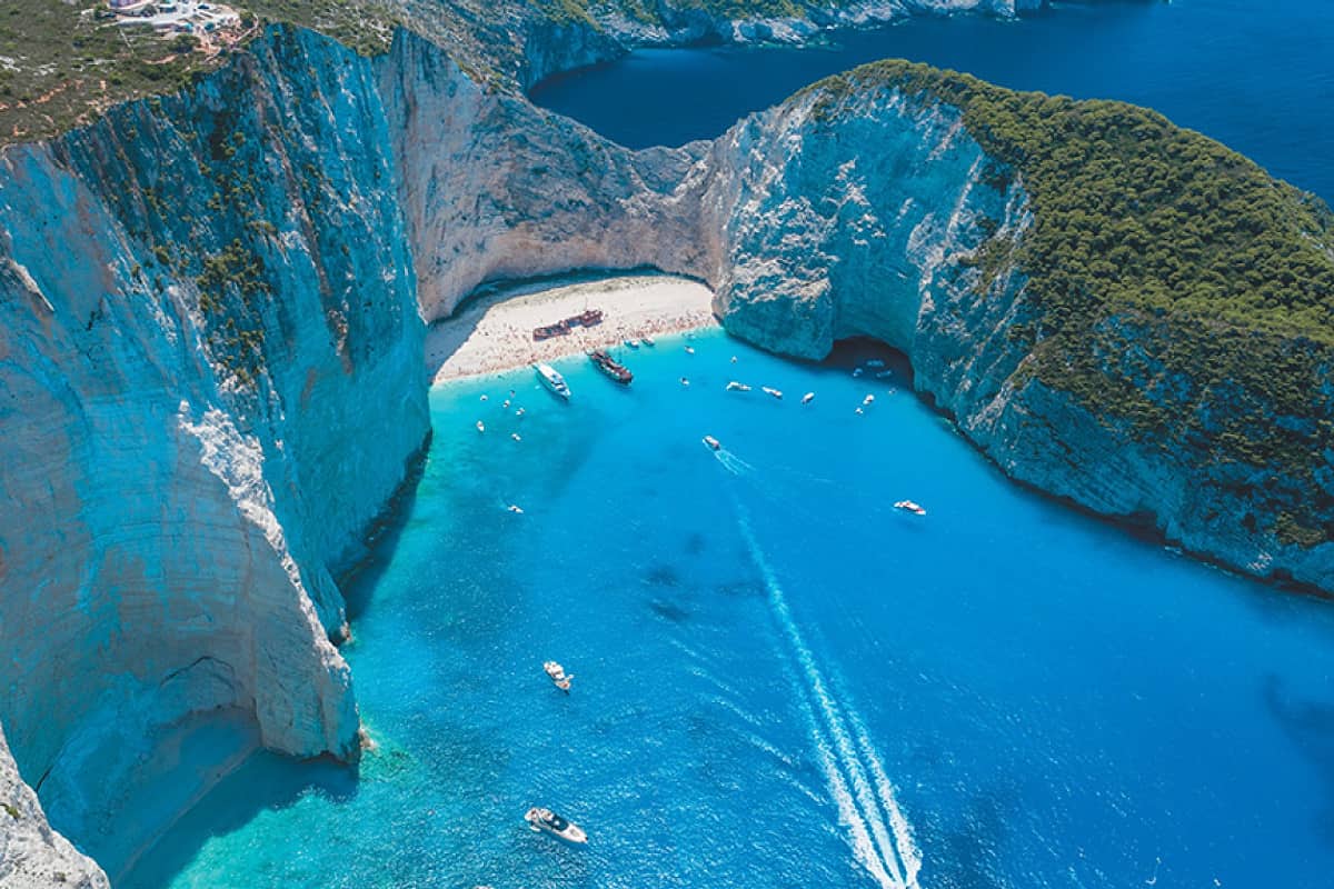 Navagio shipwreck tour boats in the turquoise cove below the white cliffs, with the rusty wreck on the beach, seen from above