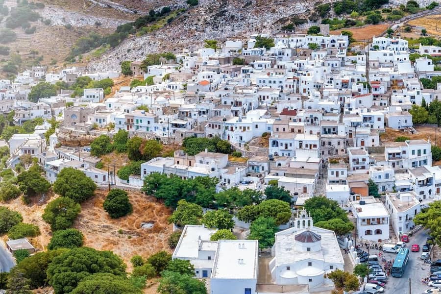Naxos day trip: an aerial view of a mountain village of white flat-roofed houses climbing a rocky slope, with a white church with a domed roof and a small bell gable at the bottom and green trees around it.