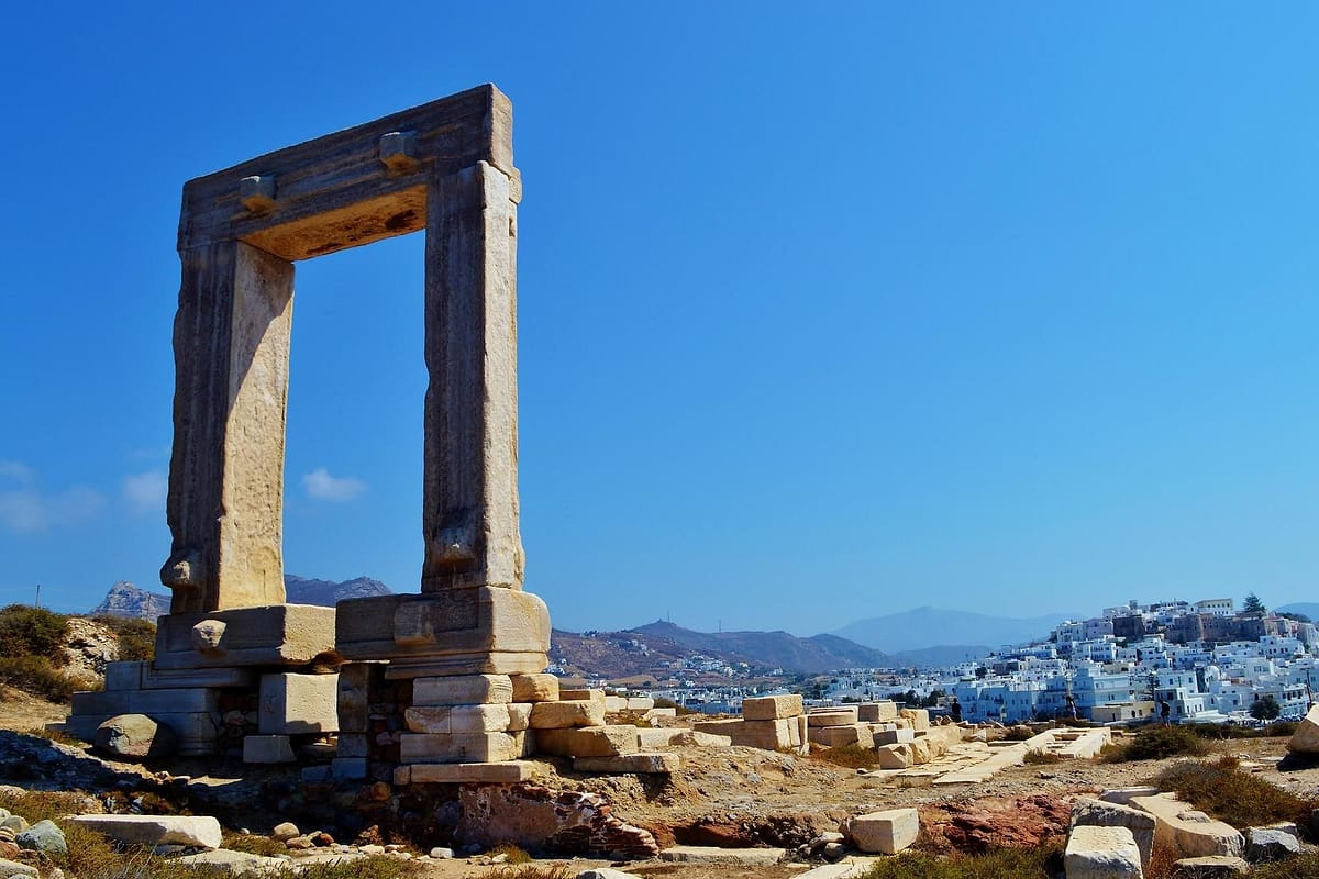 Naxos day trip: the Portara, a tall marble gateway of two upright blocks and a heavy lintel, stands on a rocky hilltop strewn with marble blocks under a deep blue sky, with the white houses of Naxos Town on a hillside and distant mountains on the right.