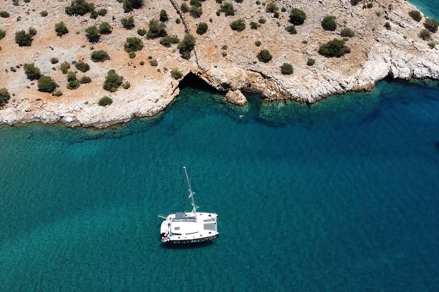 Naxos snorkeling tour: an aerial view of a white catamaran at anchor in deep turquoise water beside a rocky headland with a dark sea cave and green bushes.
