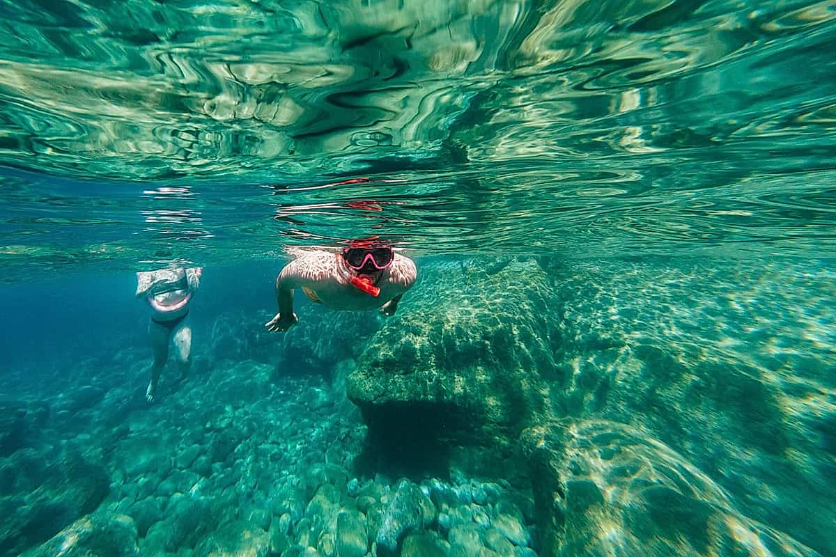 Naxos snorkeling tour: two snorkelers in masks swim at the surface of crystal-clear turquoise water off Naxos, Greece, above big rocks and pebbles on the sea floor.