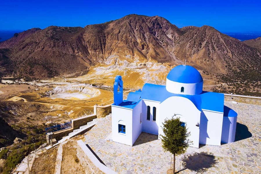 Nisyros boat trip: a white chapel with a bright blue dome and a blue bell arch stands on a paved stone terrace above the caldera of Nisyros, looking down on the pale Stefanos crater between bare brown volcanic mountains under a deep blue sky.