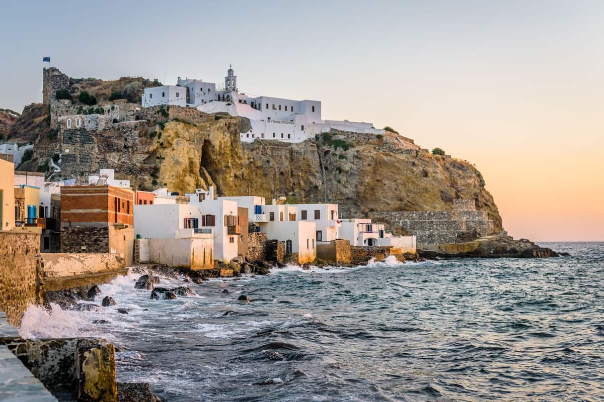 Nisyros boat trip: the white Panagia Spiliani monastery with its bell tower stands on a golden cliff above the whitewashed seaside houses of Mandraki on Nisyros, with waves breaking on the rocky shore under a pastel sunset sky.