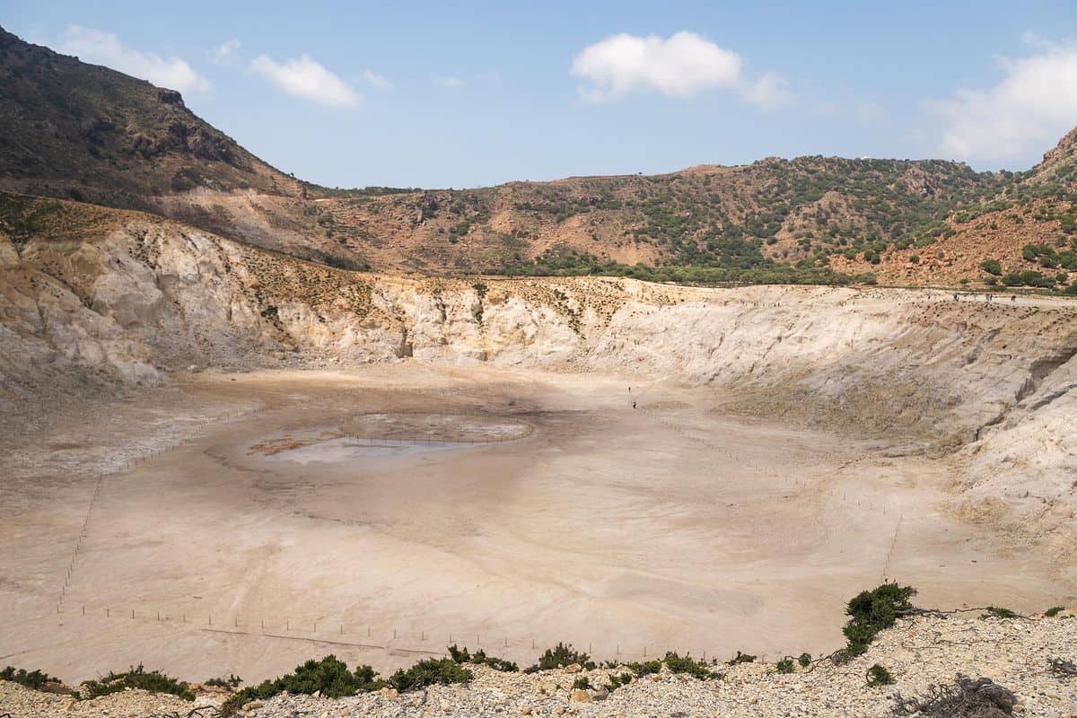 Nisyros day trip: a wide volcanic crater with a flat pale sandy floor and a small muddy pool, ringed by steep white and yellow rock walls and brown hills under a blue sky with white clouds.